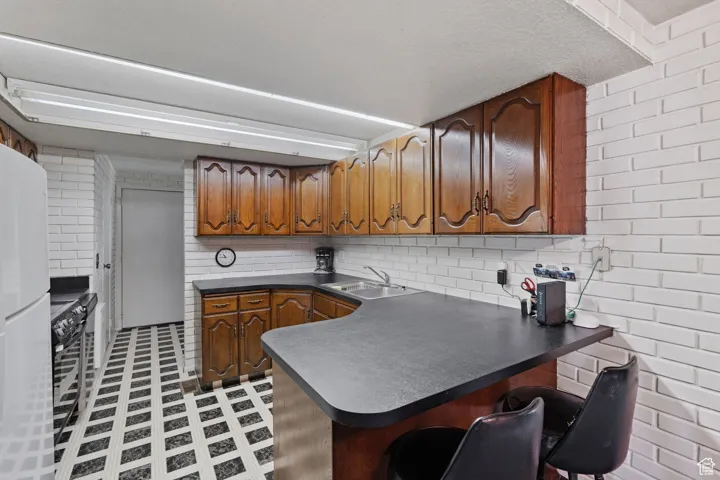 Kitchen featuring dark countertops, brown cabinets, brick wall, freestanding refrigerator, and a peninsula