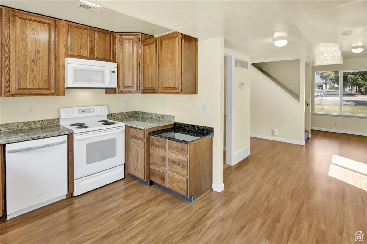 Kitchen with white appliances, brown cabinets, light wood finished floors, and dark stone countertops
