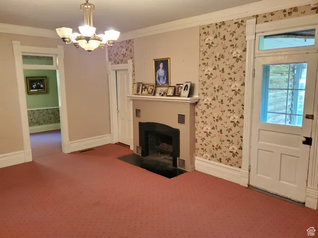 Unfurnished living room with wallpapered walls, crown molding, a fireplace with flush hearth, carpet flooring, and a chandelier