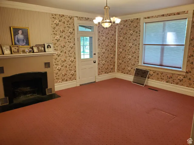 Unfurnished living room featuring wallpapered walls, ornamental molding, a chandelier, a fireplace with flush hearth, and carpet floors