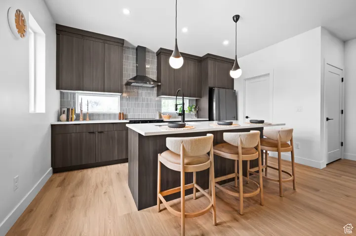 Kitchen featuring modern cabinets, a breakfast bar, an island with sink, light wood-style flooring, and wall chimney exhaust hood