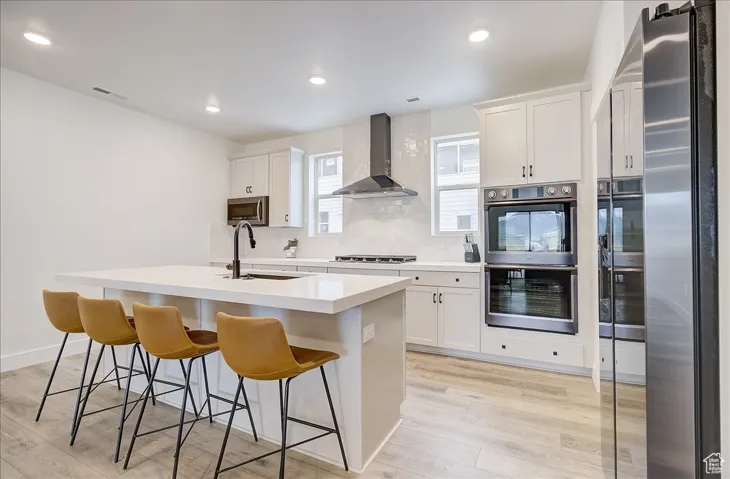 Kitchen featuring tasteful backsplash, appliances with stainless steel finishes, light wood-style flooring, white cabinetry, and wall chimney range hood