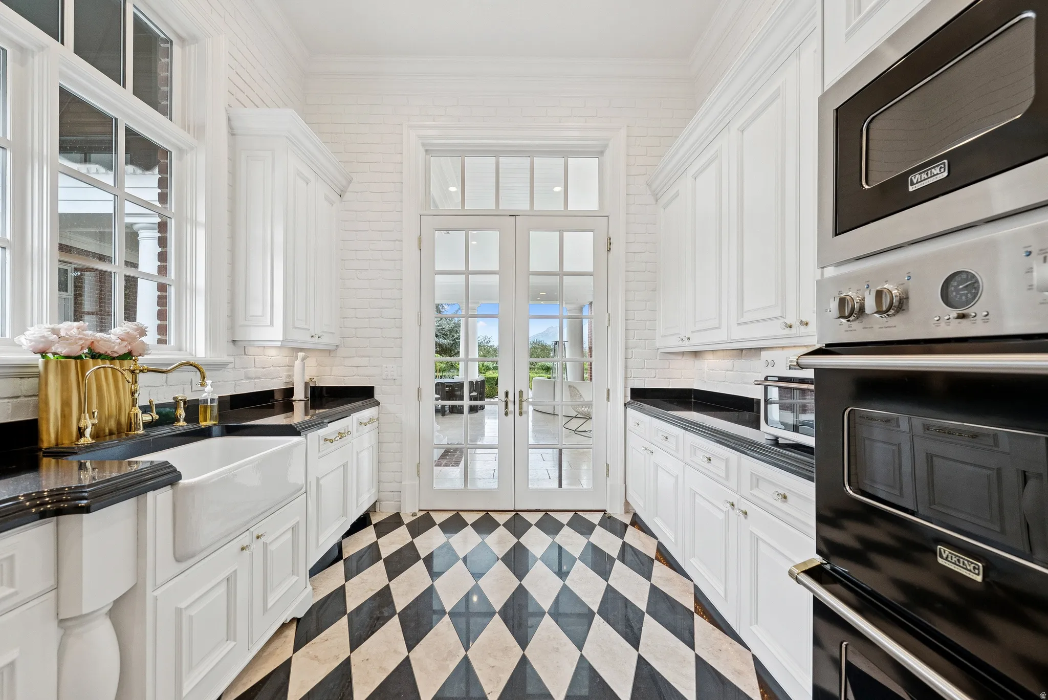 Kitchen featuring black and white diamond-patterned flooring, white shaker-style cabinetry, black countertops, and a farmhouse sink with a brass-finish faucet