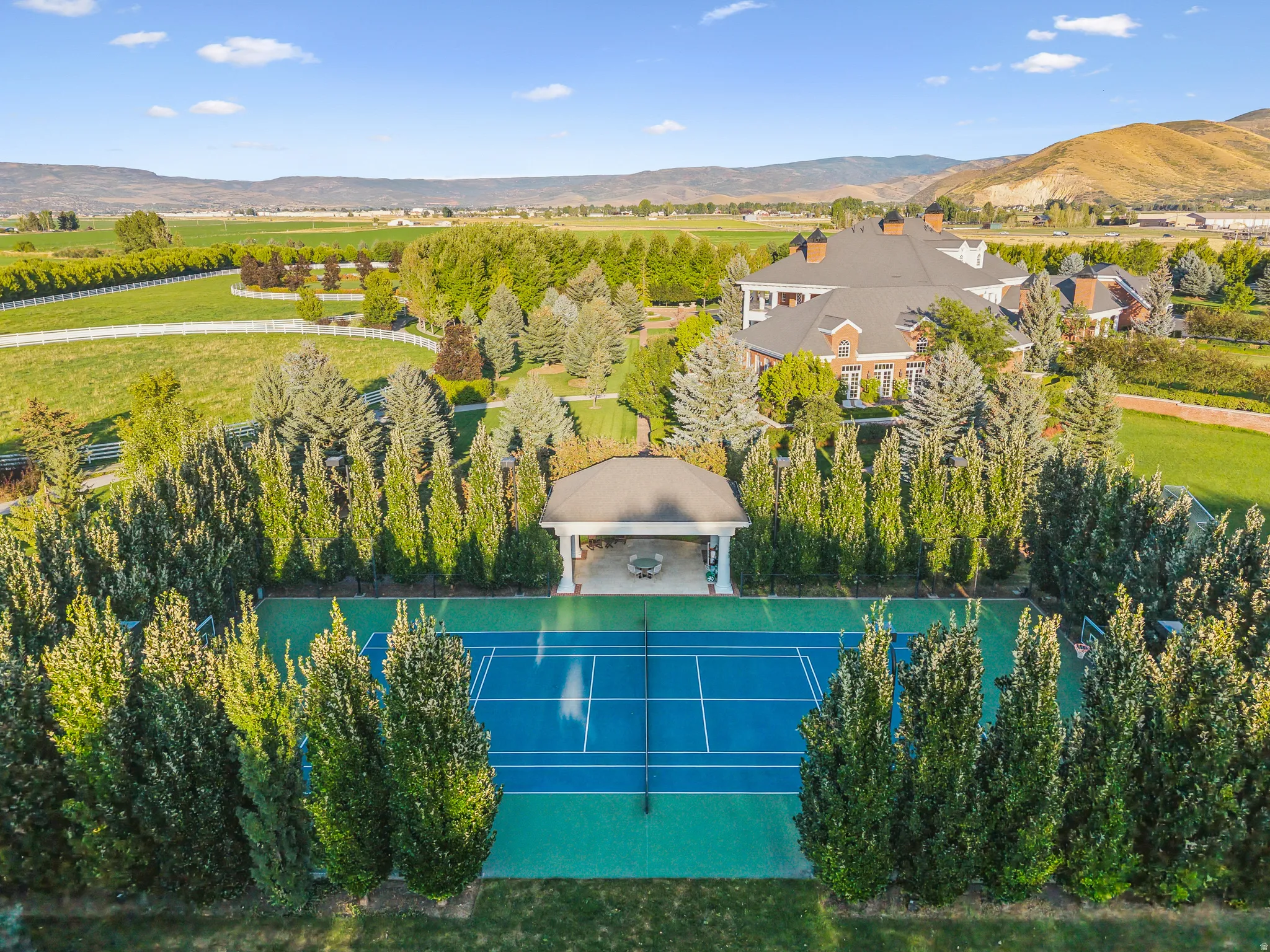 Full-size tennis court with a blue playing surface and green surrounds, bordered by mature evergreen trees