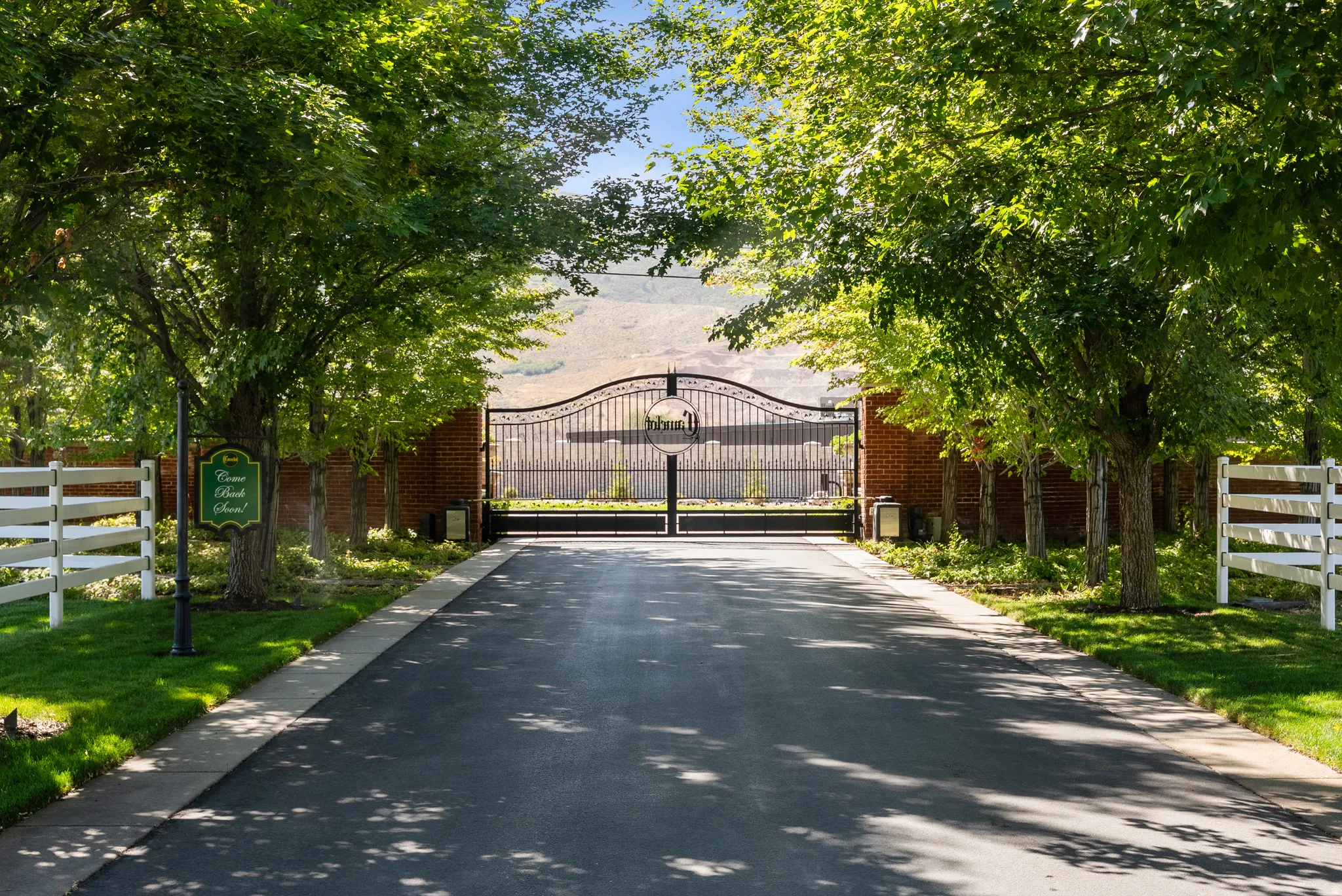 Paved driveway entry featuring an ornate wrought iron gate, brick privacy wall, white split-rail fencing, mature trees, and manicured lawns