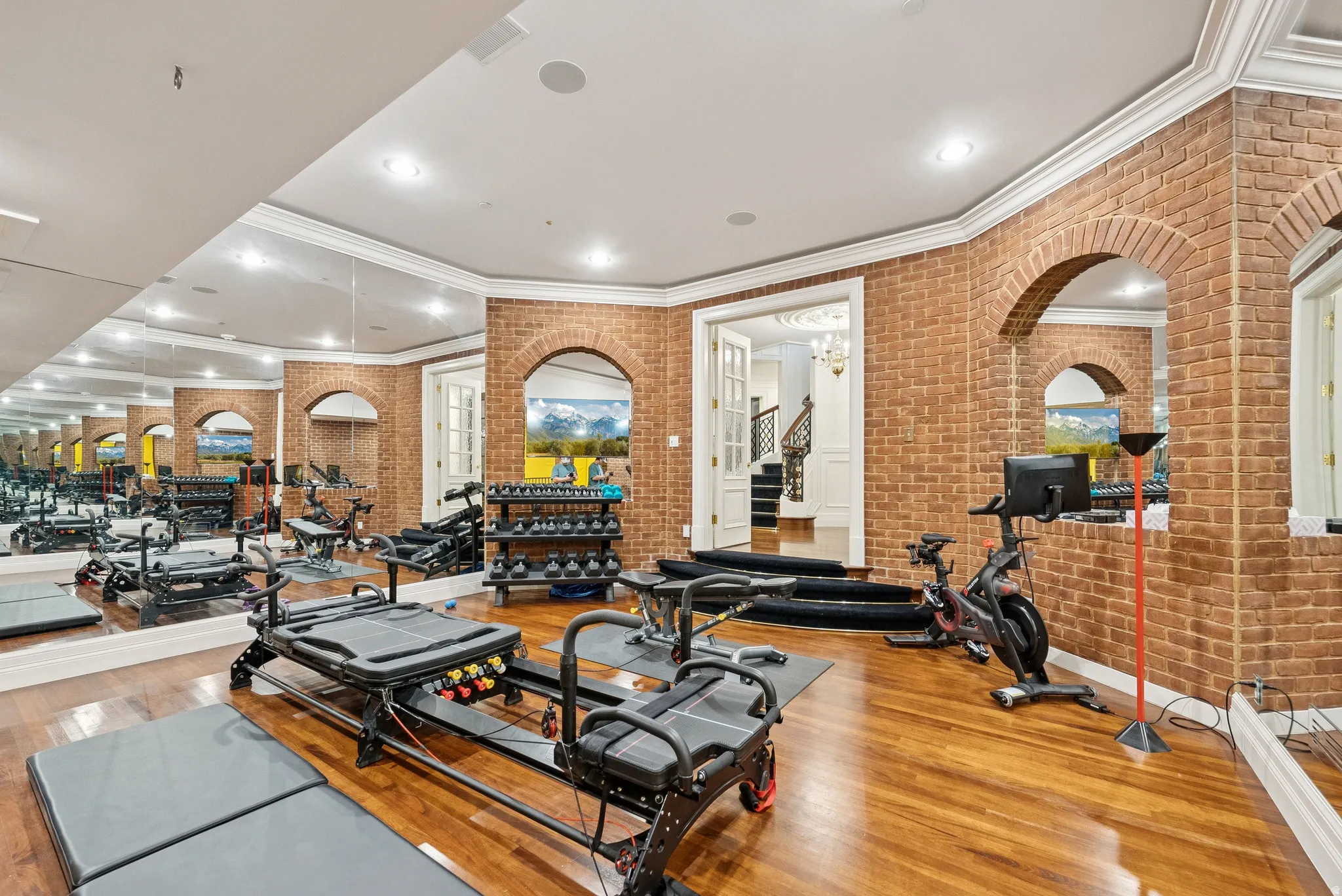 Dedicated fitness room featuring wood-finish flooring, full-wall mirrors, brick-finish walls with arched openings, and crown molding