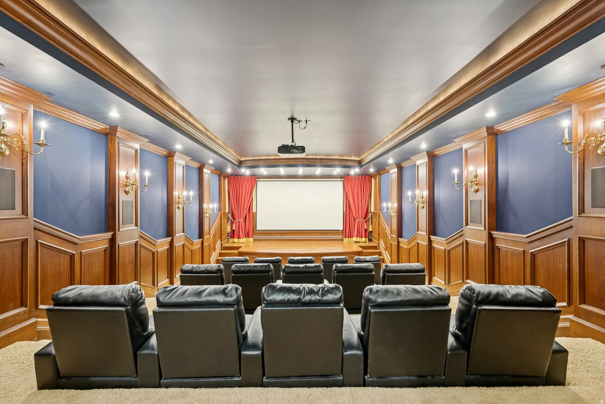 Dedicated home theater featuring tiered seating, wood paneling, sconce lighting, a projection screen, and red stage drapes