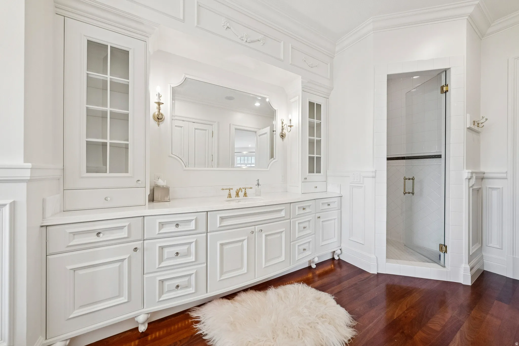 White custom cabinetry with glass-front display cabinets, a recessed vanity mirror, and dual wall sconces