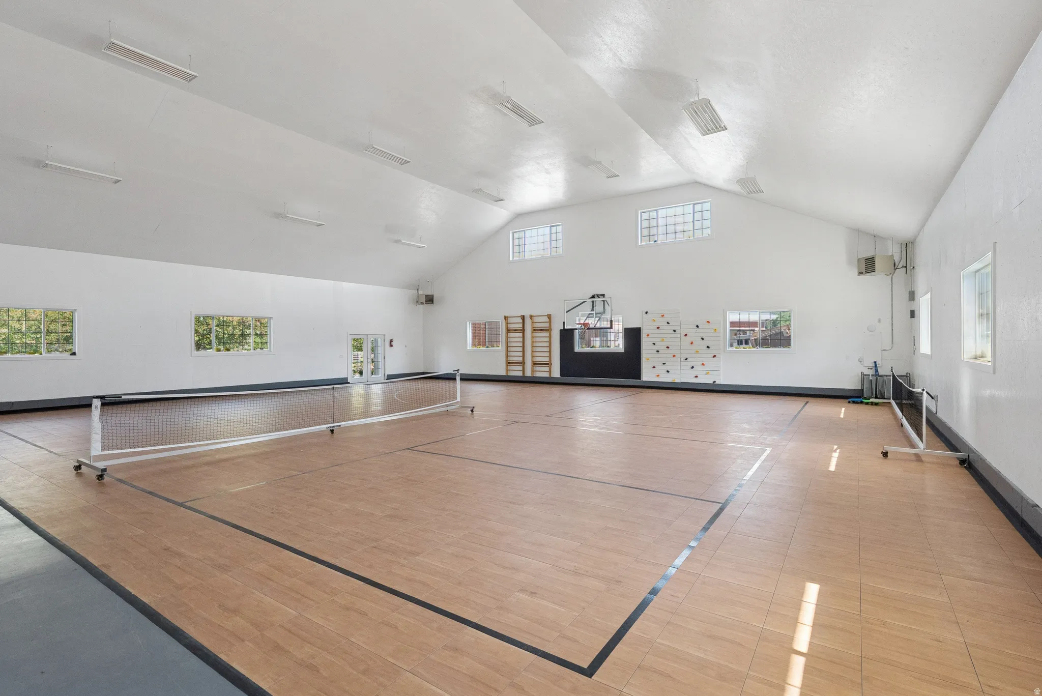 Spacious interior athletic court featuring wood-finish flooring with court lines, a full-height ceiling, multiple windows, and recessed lighting