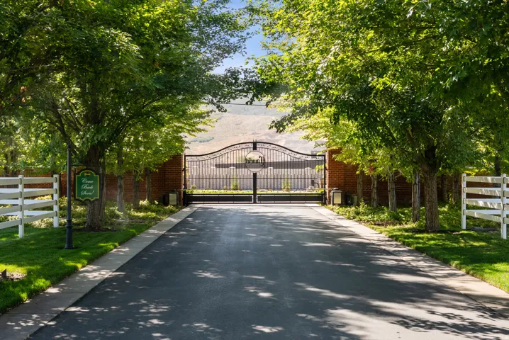 Paved driveway entry featuring an ornate wrought iron gate, brick privacy wall, white split-rail fencing, mature trees, and manicured lawns