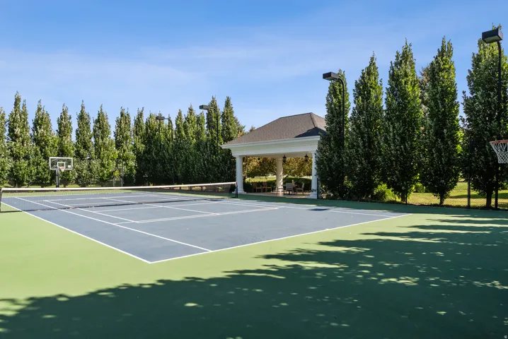 Multi-sport court featuring a tennis net, basketball hoops, and a perimeter of mature trees