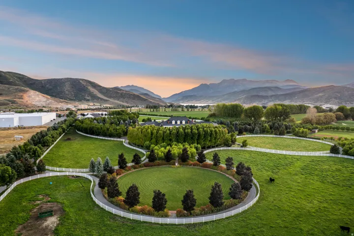Expansive property featuring manicured green pastures, white fencing, mature trees, a circular driveway, and mountainous scenery