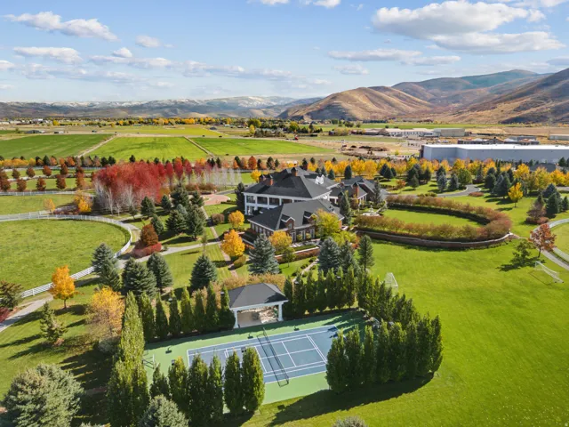 Expansive estate featuring a large main residence, a tennis court with a shaded pavilion, and a surrounding landscape of mature trees