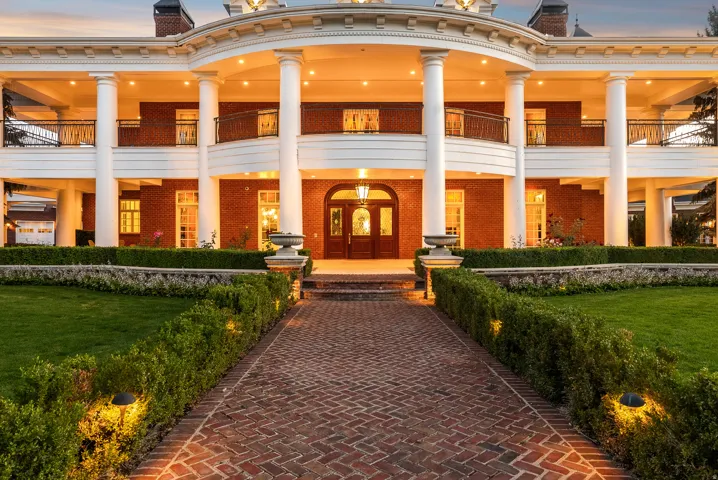 Stately brick residence featuring a grand entrance with a columned portico, a curved second-story balcony with metal railings, and a meticulously landscaped front yard