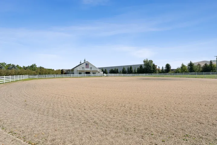 Expansive outdoor riding arena with a white perimeter fence, featuring a large white barn structure in the background
