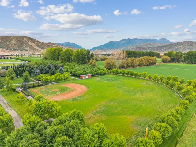 Expansive green field featuring a baseball diamond, surrounded by mature trees, a red and white storage shed, and a mountain backdrop