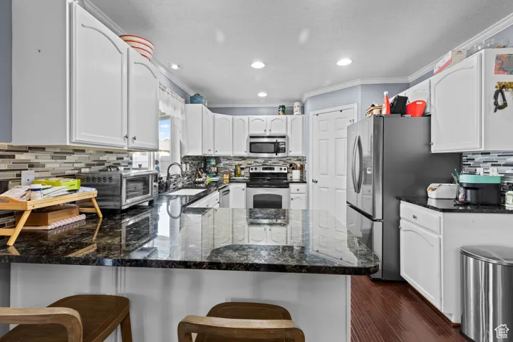 Kitchen with a peninsula, white cabinets, ornamental molding, appliances with stainless steel finishes, and dark stone countertops