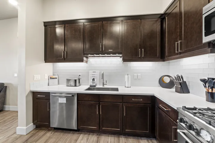 Kitchen with dark brown cabinetry, stainless steel appliances, tasteful backsplash, light wood-style floors, and light stone counters