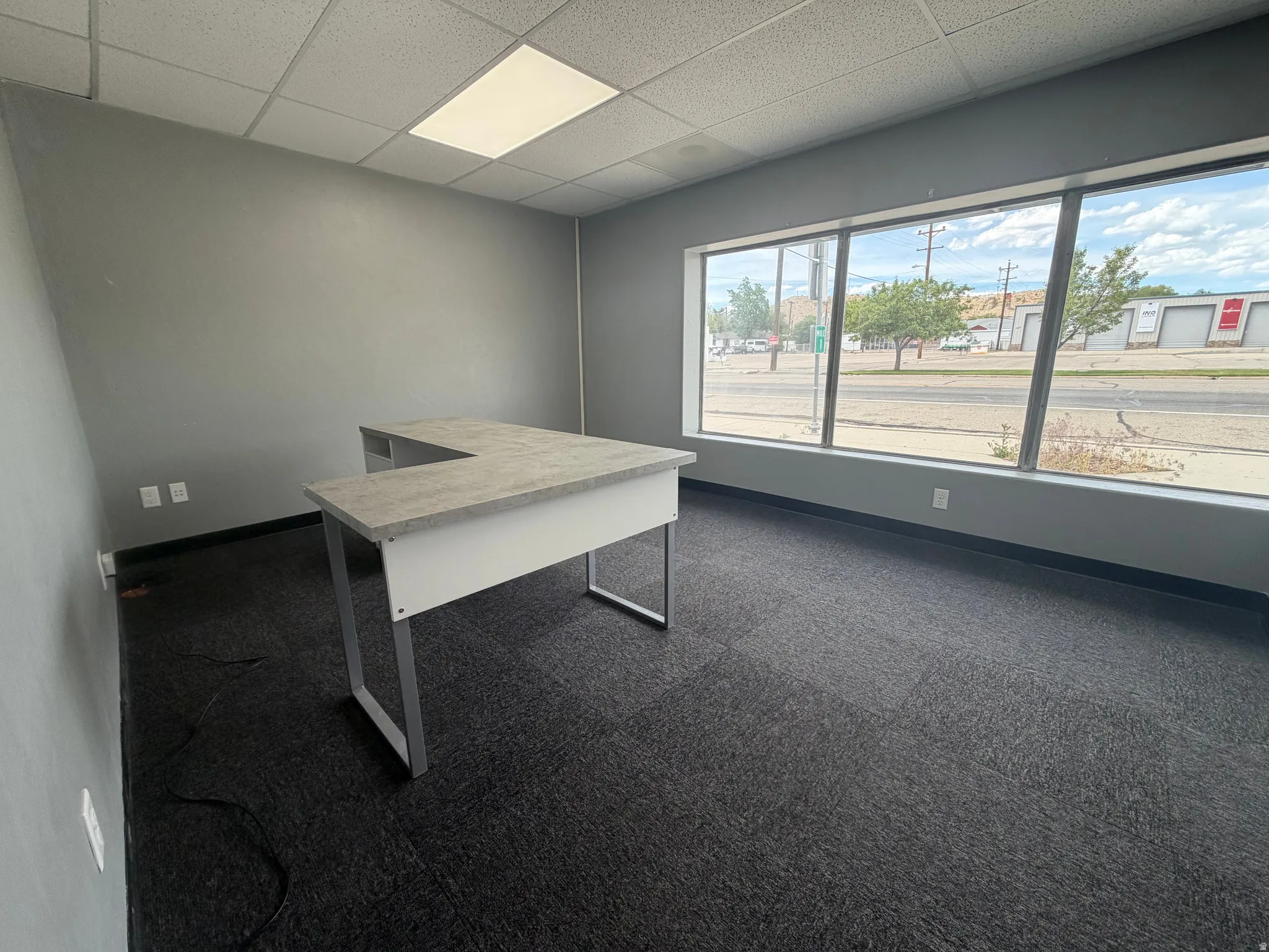 Office space featuring full-length windows, dark grey carpet flooring, light grey painted walls, and a dropped ceiling with recessed lighting