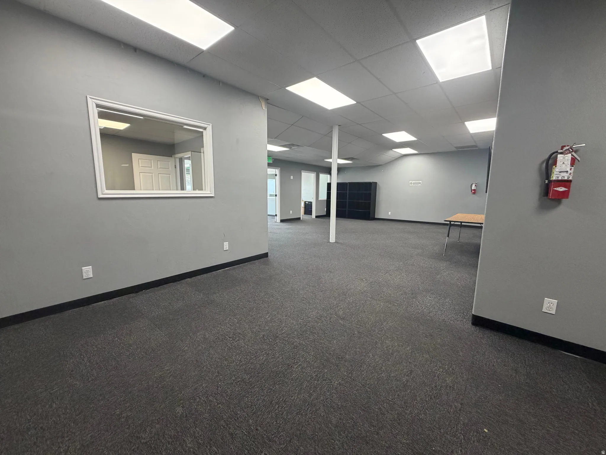 Open-concept commercial interior featuring gray walls, dark patterned carpet, and a drop ceiling with integrated rectangular lighting