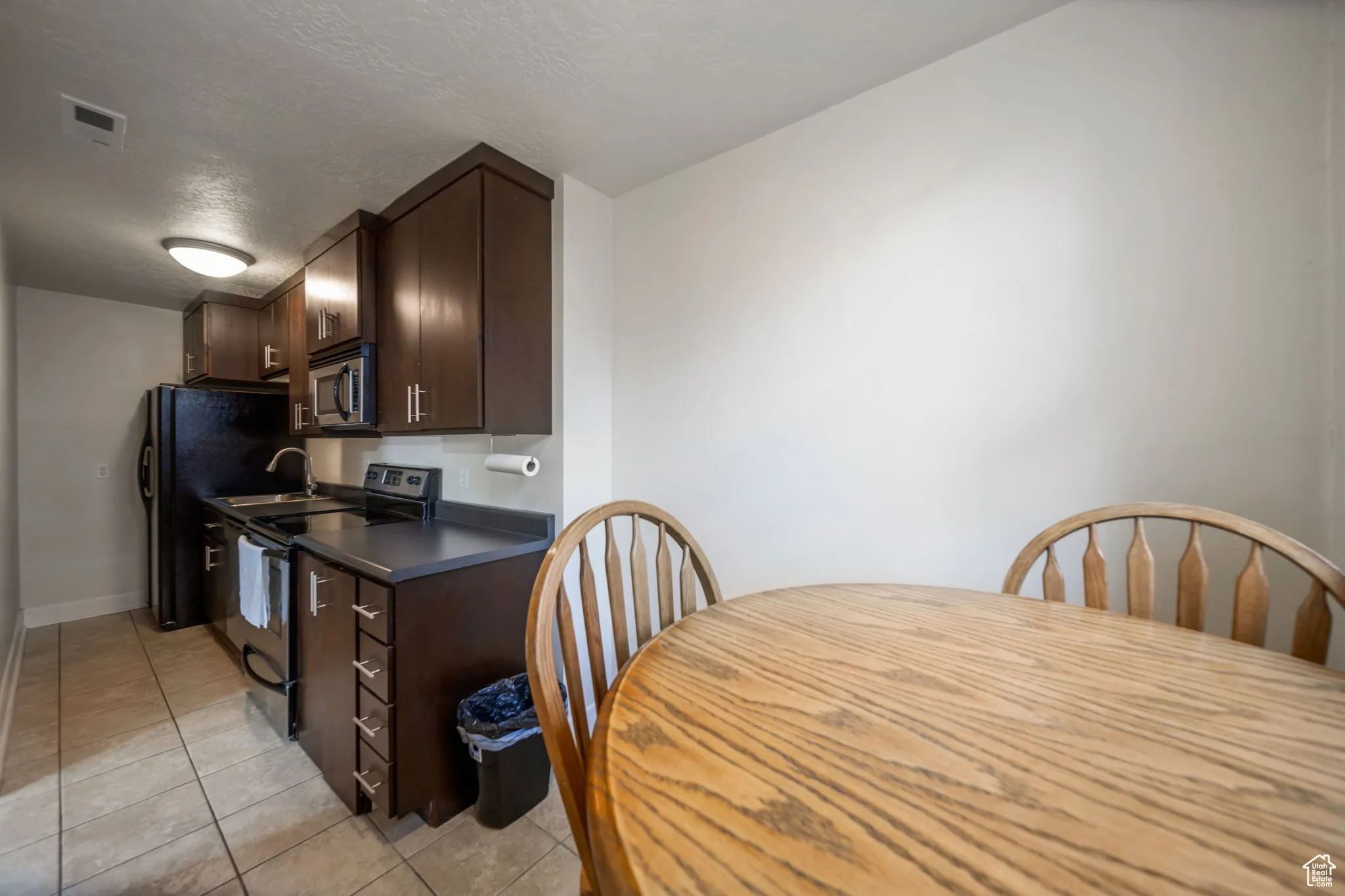Kitchen featuring dark brown cabinets, stainless steel appliances, dark countertops, light tile patterned floors, and a textured ceiling
