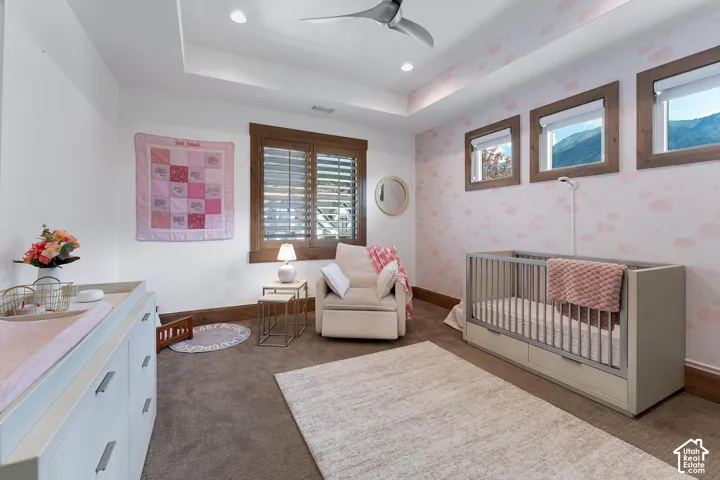 Bedroom featuring a crib, a raised ceiling, dark carpet, wallpapered walls, and ceiling fan