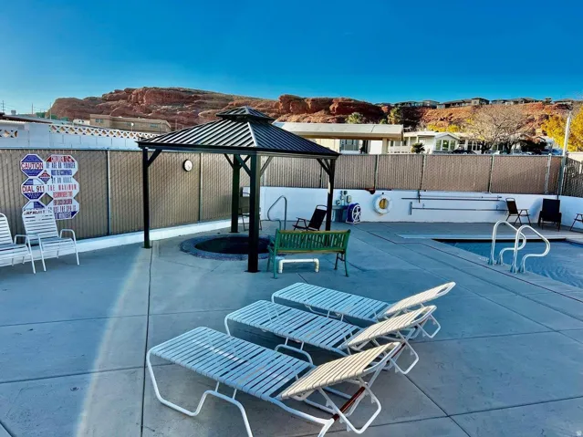 Resort-style pool deck featuring multiple lounge chairs, a black metal pergola with integrated shade structure, concrete decking, and scenic red rock backdrop