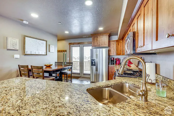 Kitchen featuring appliances with stainless steel finishes, brown cabinets, a textured ceiling, light stone countertops, and recessed lighting