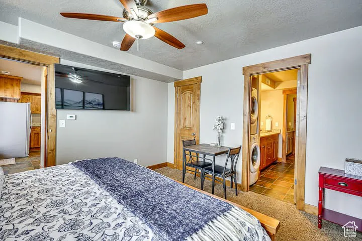 Bedroom featuring ensuite bath, freestanding refrigerator, a textured ceiling, a ceiling fan, and stacked washer / dryer