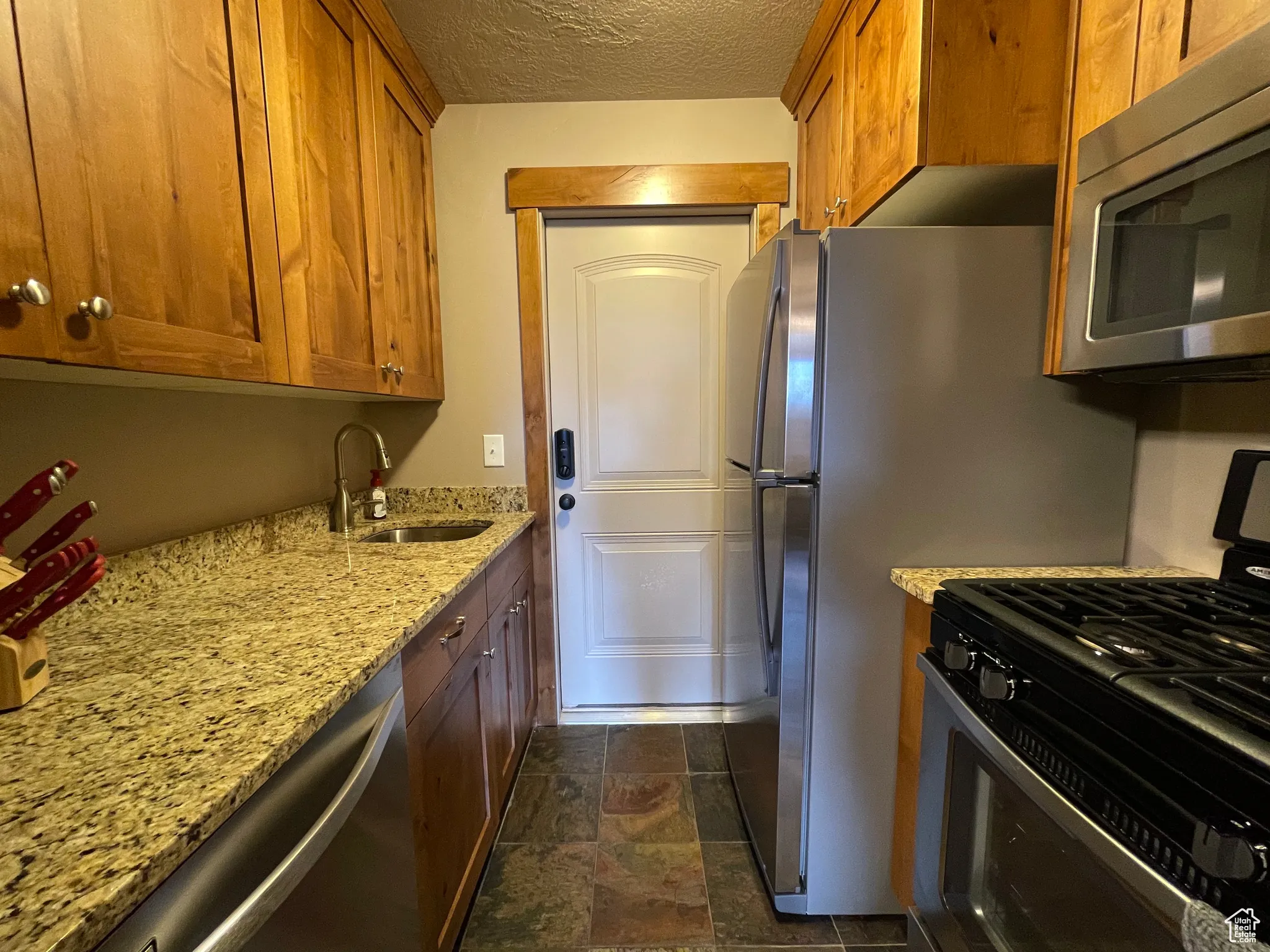 2nd Kitchen featuring appliances with stainless steel finishes, a textured ceiling, light stone countertops, brown cabinetry, and dark stone finish floors