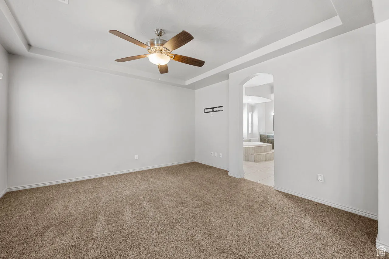 Empty room with a raised ceiling, light carpet, arched walkways, and ceiling fan