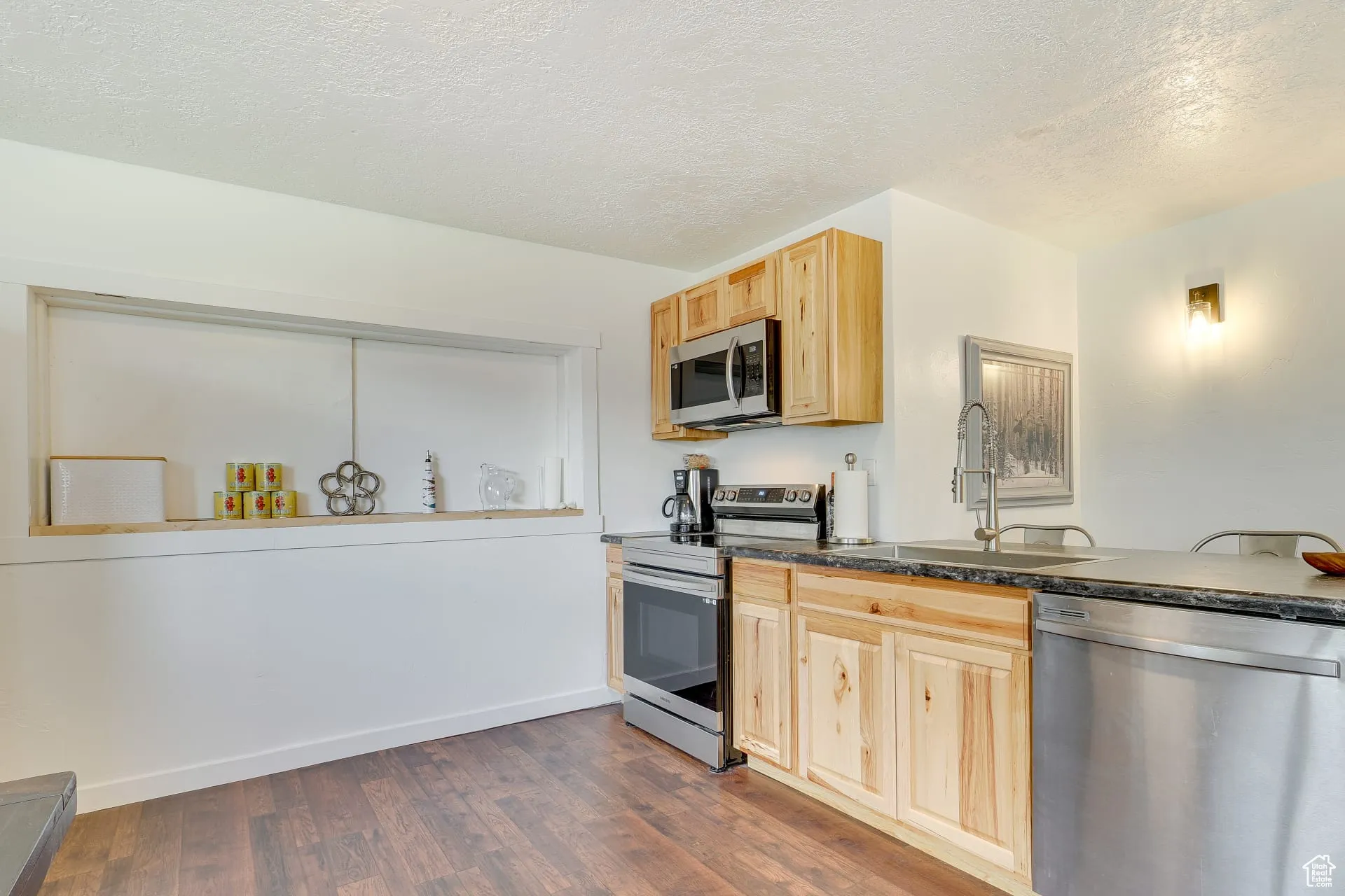 Kitchen featuring a textured ceiling, appliances with stainless steel finishes, light brown cabinetry, dark wood-type flooring, and dark stone counters