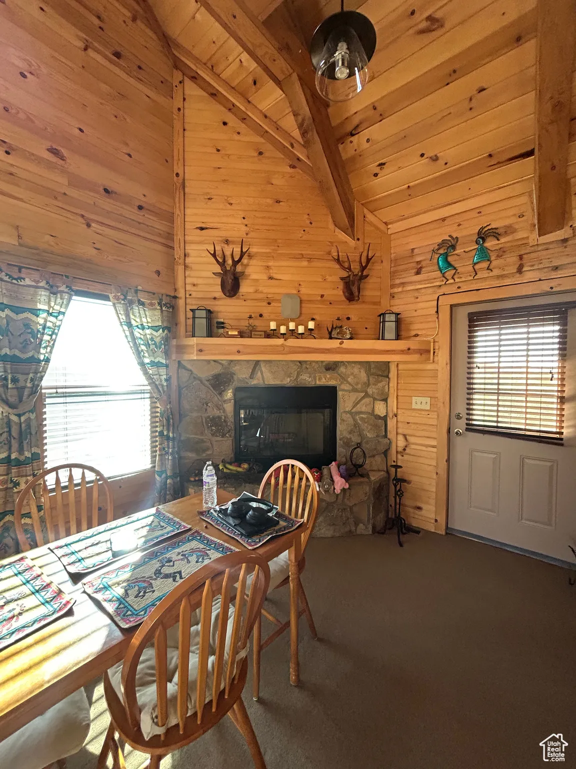 Carpeted dining area with wood walls, a stone fireplace, a wooden ceiling with exposed beams, and high vaulted ceiling