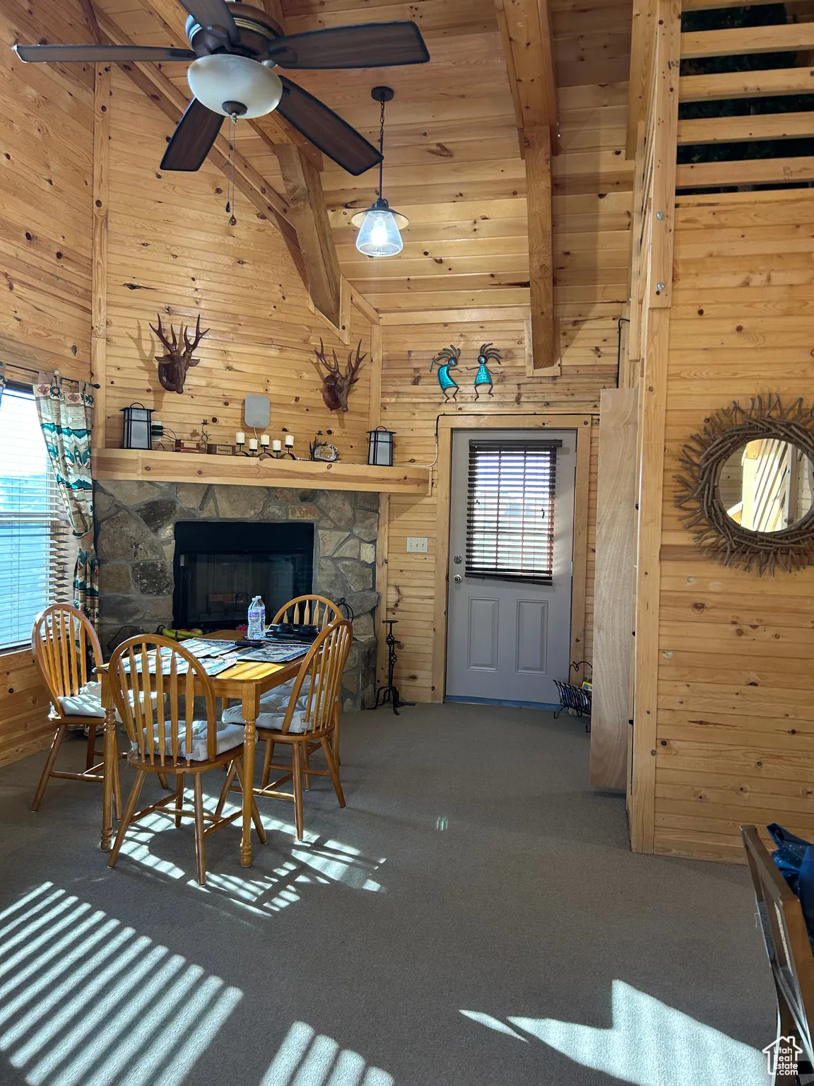 Carpeted dining room featuring wooden walls, a stone fireplace, high vaulted ceiling, a wood ceiling with exposed beams, and a ceiling fan