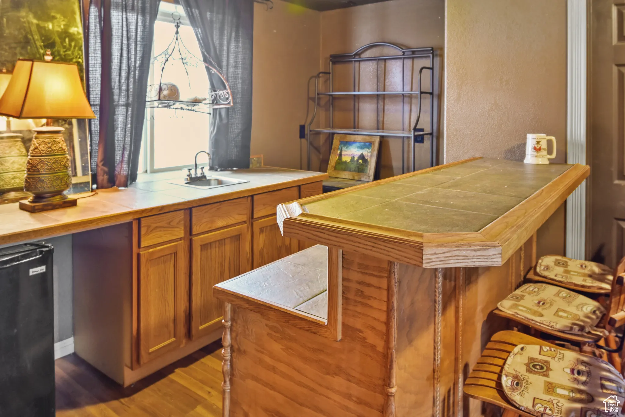 Bar with brown cabinetry, black refrigerator, light wood-type flooring, tile counters, and a textured wall
