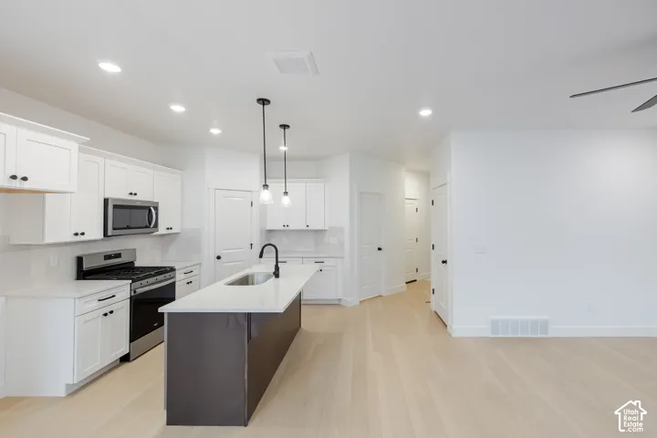 Modern kitchen featuring white cabinetry, stainless steel appliances, a center island with a sink, pendant lighting, and light wood-finish flooring