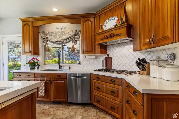 Kitchen with brown cabinets, decorative backsplash, appliances with stainless steel finishes, light stone counters, and recessed lighting