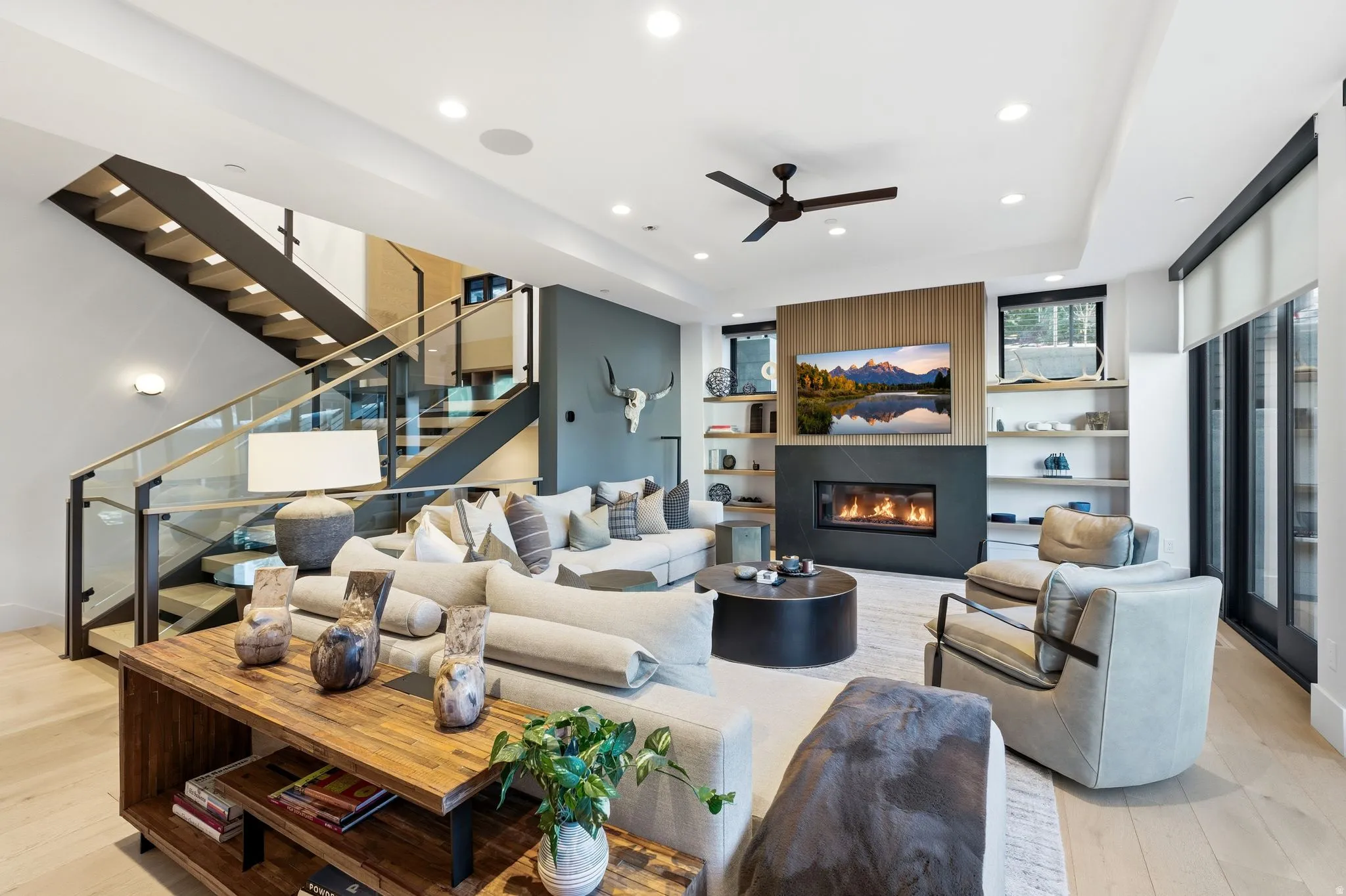 Living room with built in shelves, light wood-type flooring, a ceiling fan, a glass covered fireplace, and recessed lighting