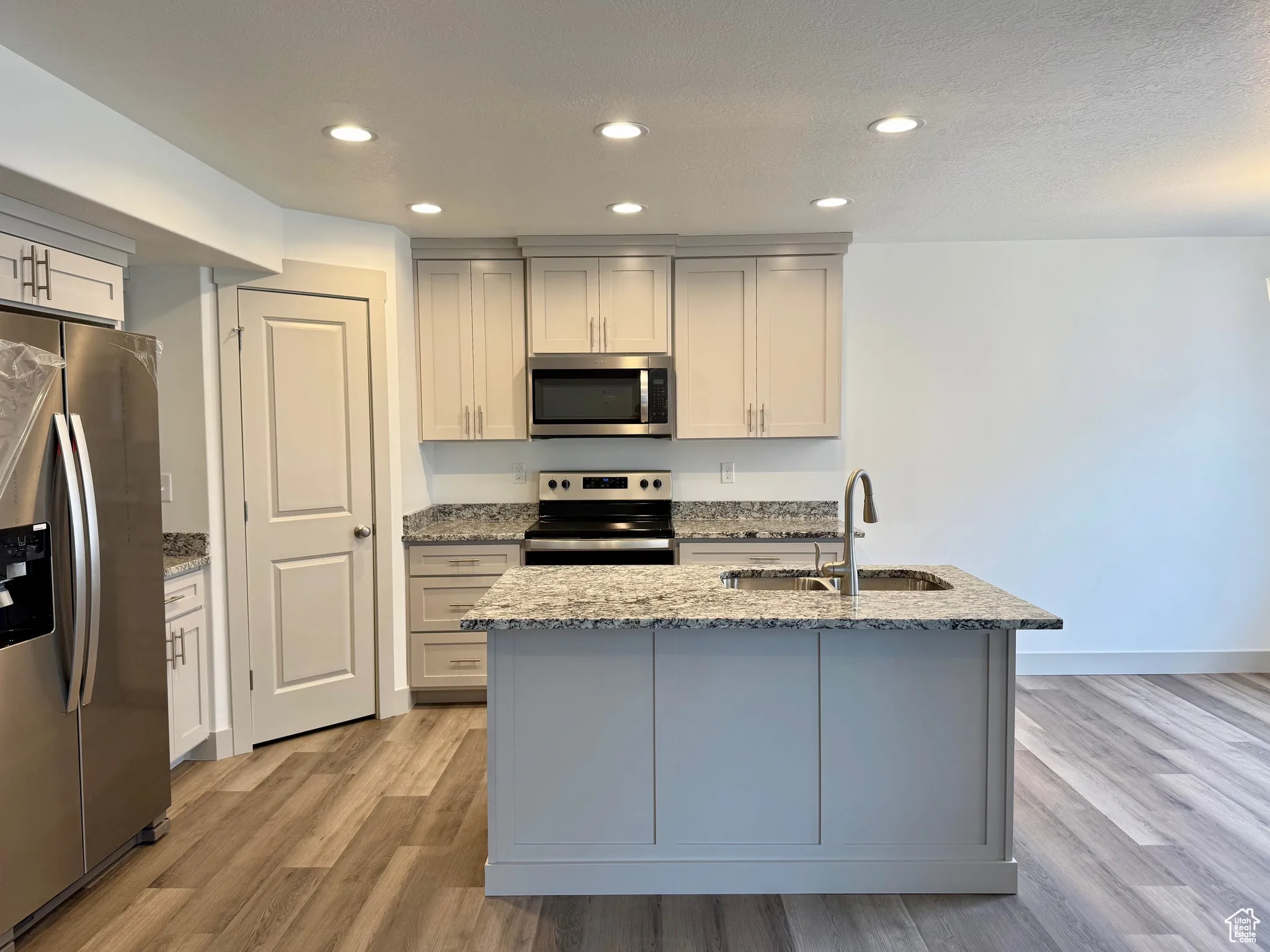 Kitchen with light stone countertops, appliances with stainless steel finishes, recessed lighting, light wood-style flooring, and an island with sink