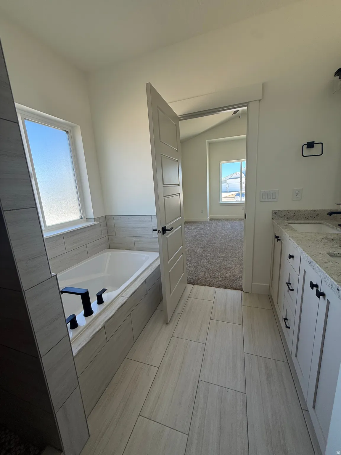 Built-in bathtub with black fixtures, light-toned tile flooring, white vanity with stone countertop, and white cabinetry with black hardware