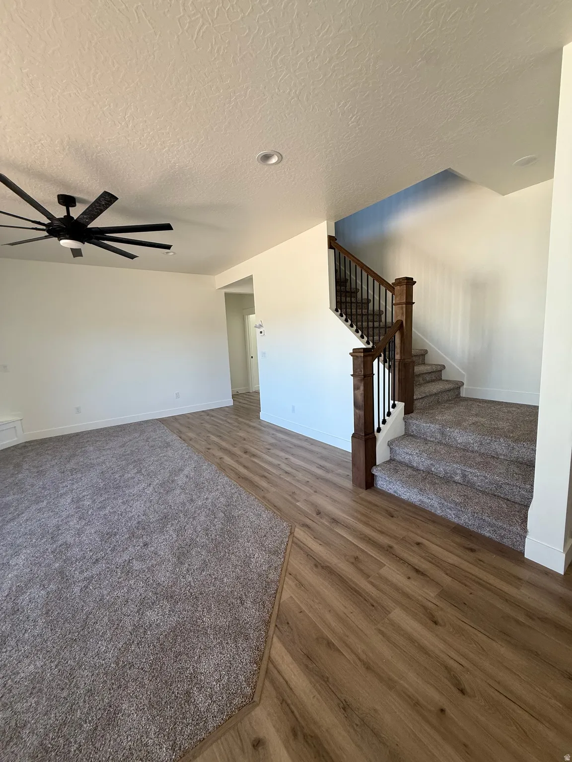 Wood-finish flooring with complementary carpeted areas, a staircase featuring wood newel posts and metal balusters, and a ceiling fan with integrated lighting