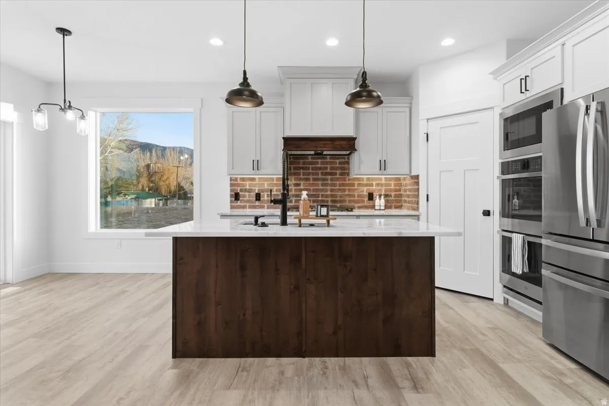 Kitchen featuring appliances with stainless steel finishes, decorative light fixtures, light stone counters, white cabinetry, and a kitchen island with sink