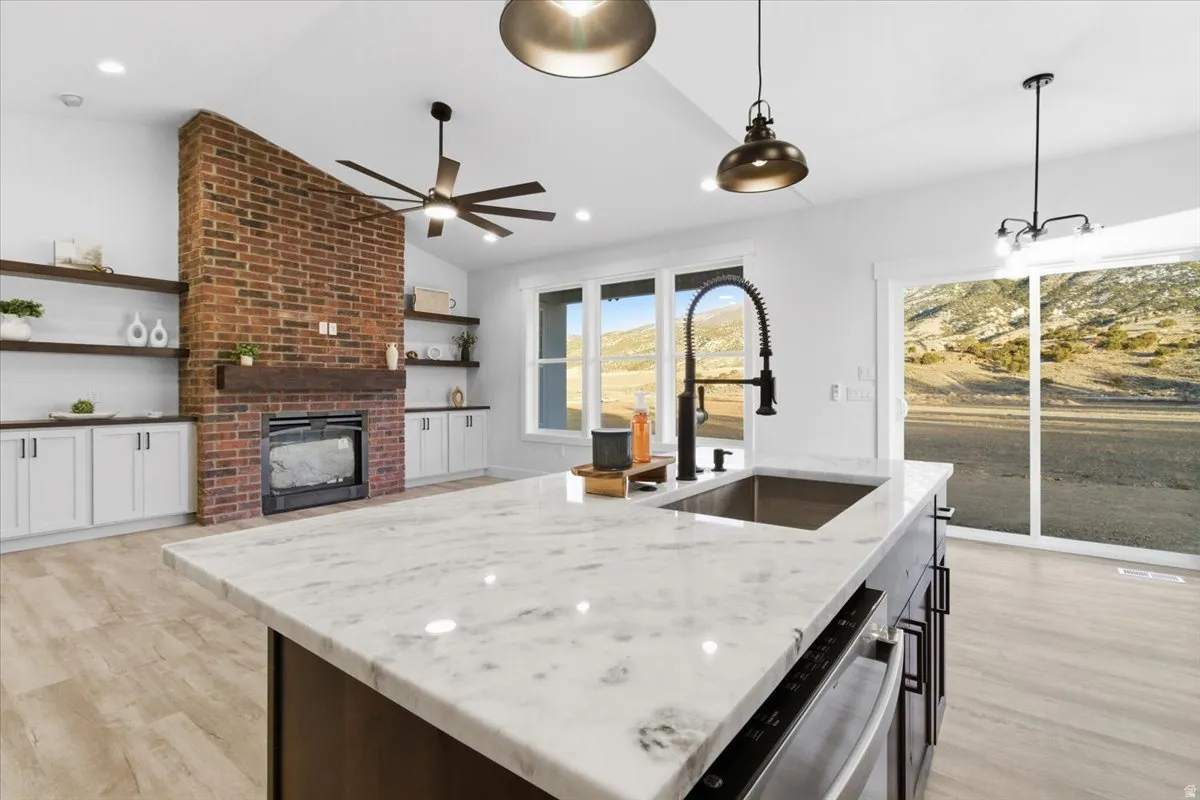 Kitchen with vaulted ceiling, decorative light fixtures, a kitchen island with sink, a brick fireplace, and light wood-type flooring