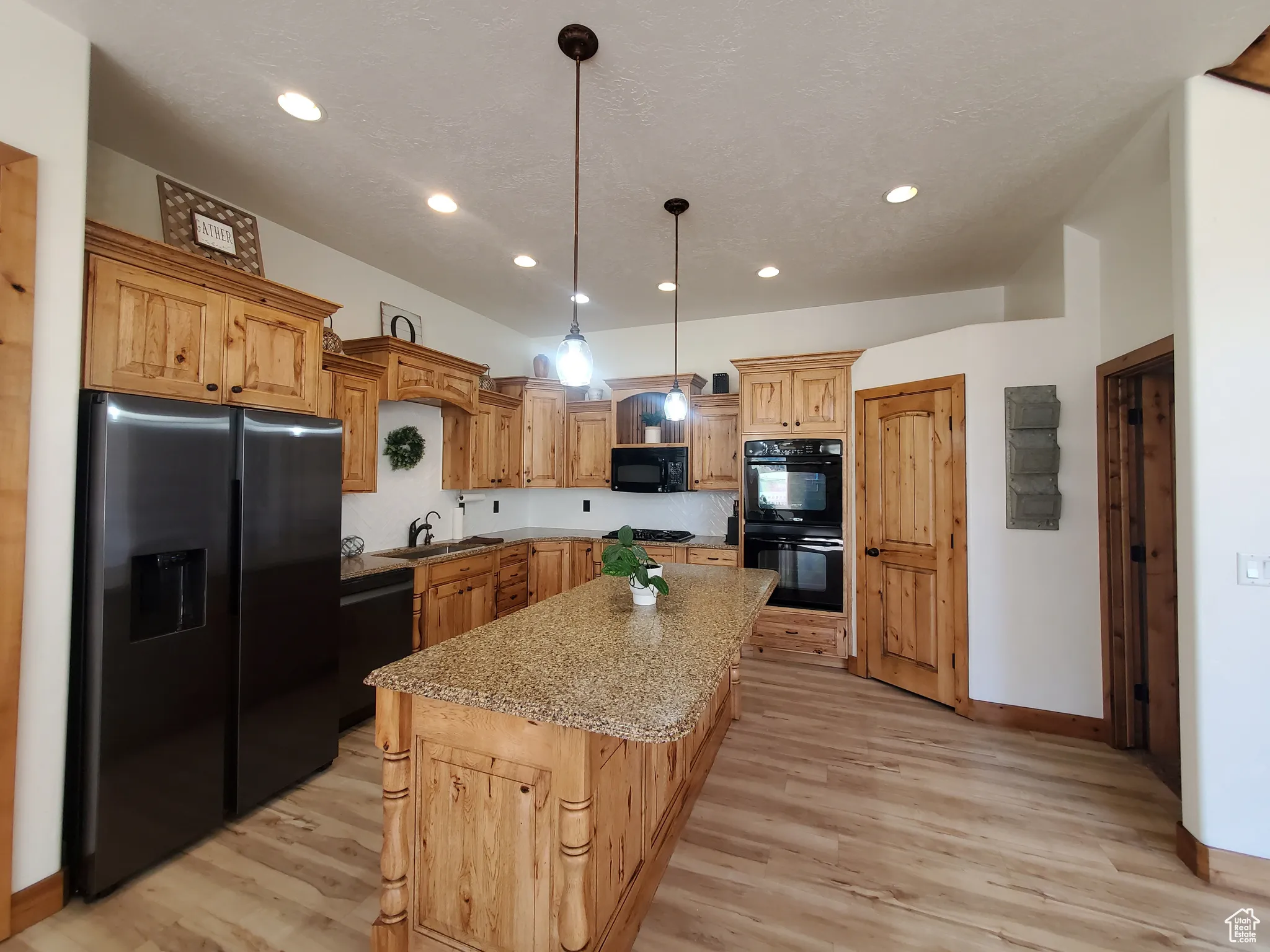 Kitchen with black appliances, light wood-type flooring, pendant lighting, light stone counters, and a center island