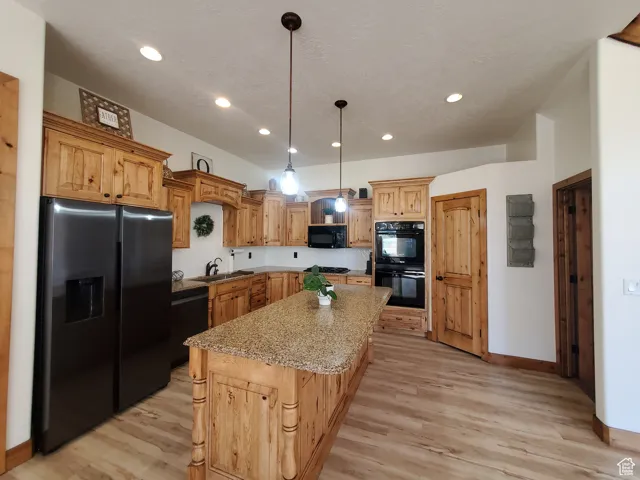 Kitchen with black appliances, light wood-type flooring, pendant lighting, light stone counters, and a center island