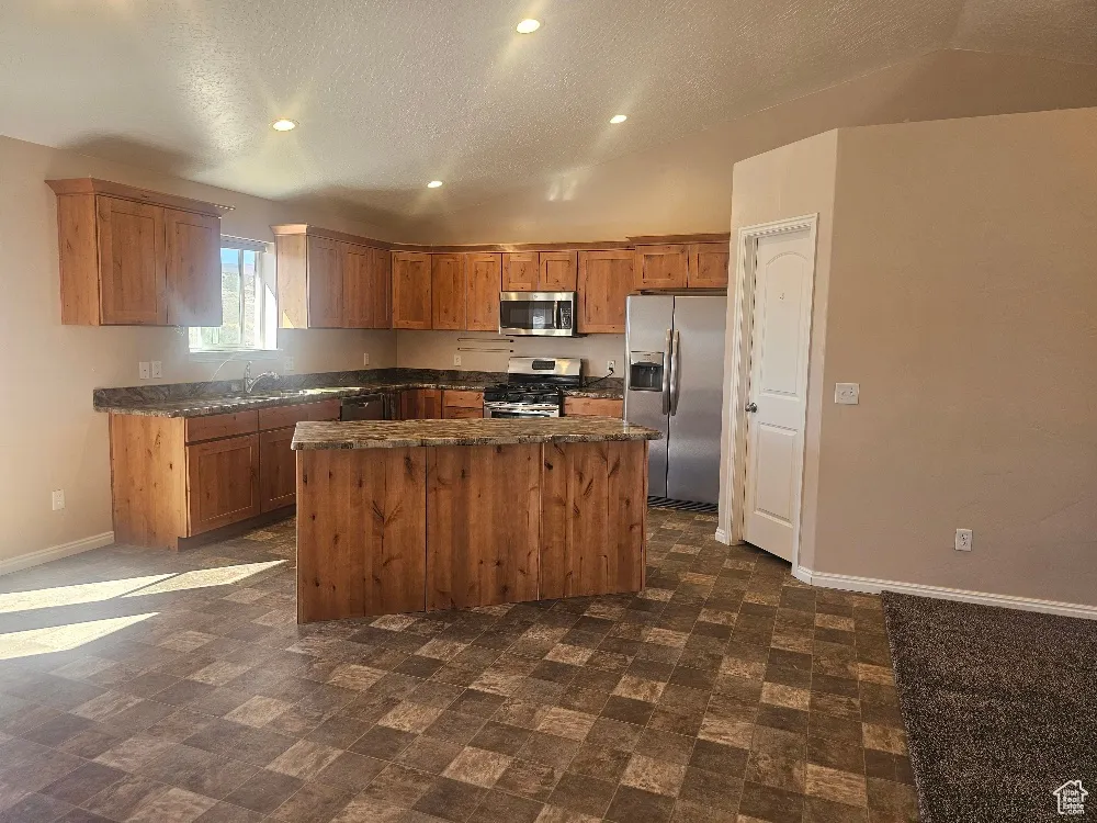 Kitchen featuring vaulted ceiling, brown cabinetry, a center island, stainless steel appliances, and dark stone countertops