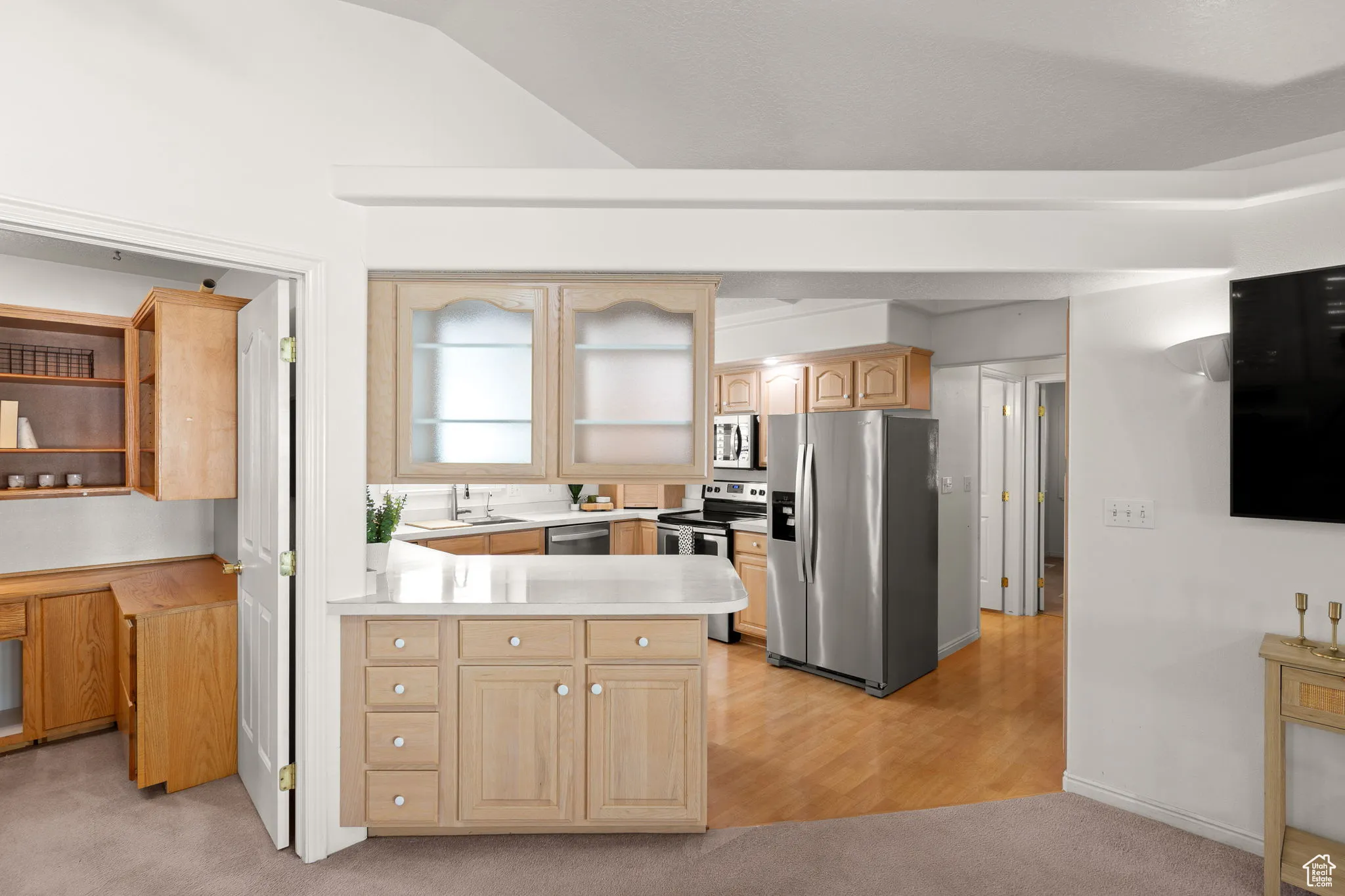 Kitchen with stainless steel appliances, a peninsula, light brown cabinets, light wood-type flooring, and light colored carpet