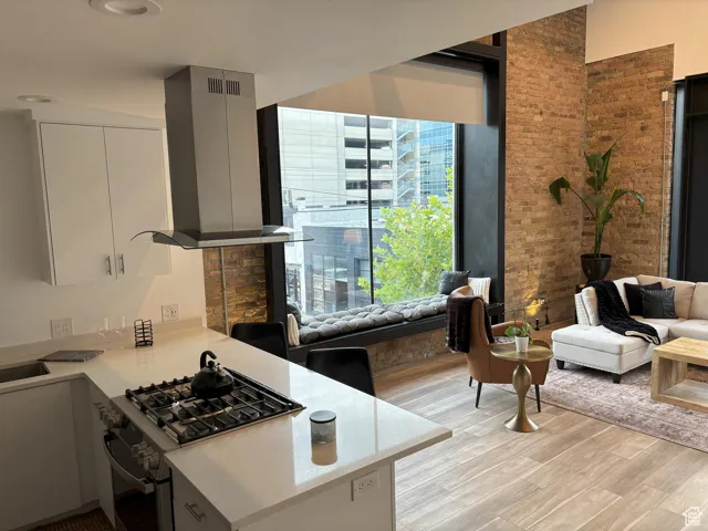 Kitchen with stainless steel range with gas cooktop, wall chimney range hood, light wood-style floors, light stone countertops, and brick wall
