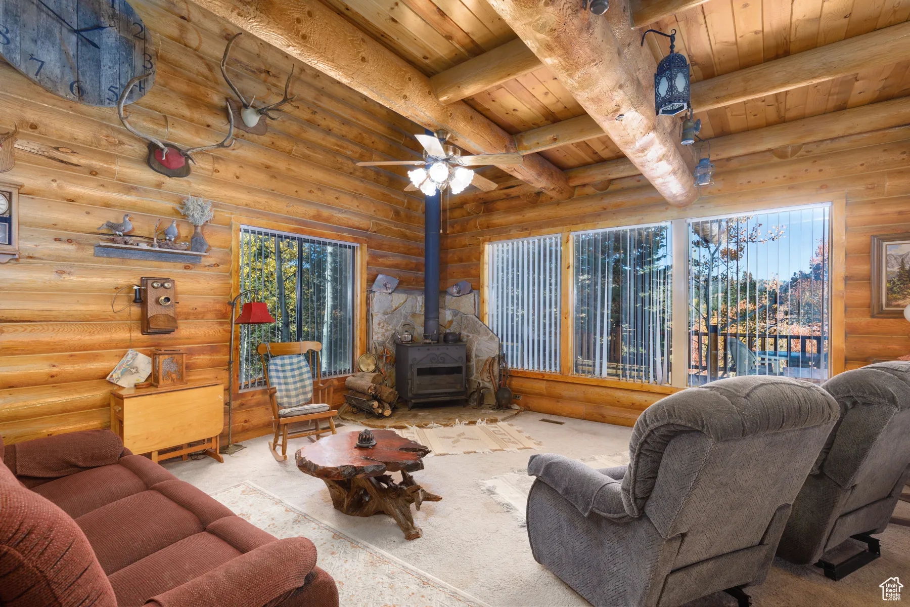 Carpeted living room with a wood stove, rustic walls, a ceiling fan, and a wood ceiling with exposed beams