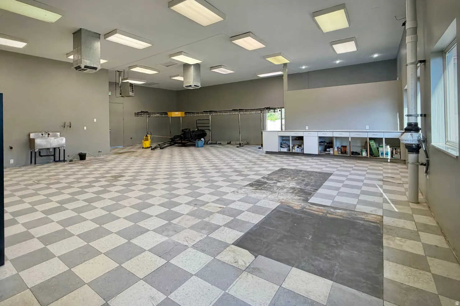 Spacious utility area featuring checkered flooring, industrial fluorescent lighting, high ceilings, built-in cabinetry, and a large utility sink